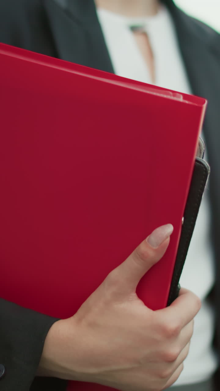 Partial view of secretary in black suit holding file folder and coffee cup while walking along path with blurred background of parked cars and urban buildings