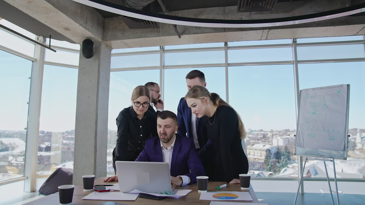 Group of colleagues stand around the boss with a laptop. Other businessman has a telephone conversation. Team working in a spacious office.