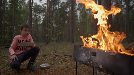 un chico adolescente guapo comiendo comida rápida cerca de una hoguera de barbacoa en el bosque.