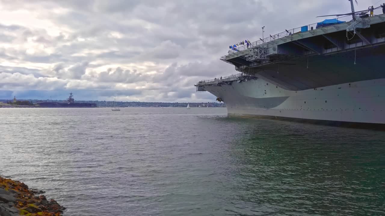 Boats floating on San Diego bay near a big navy and Tuna Harbor Park in daylight. Cloudy sky. Slow motion