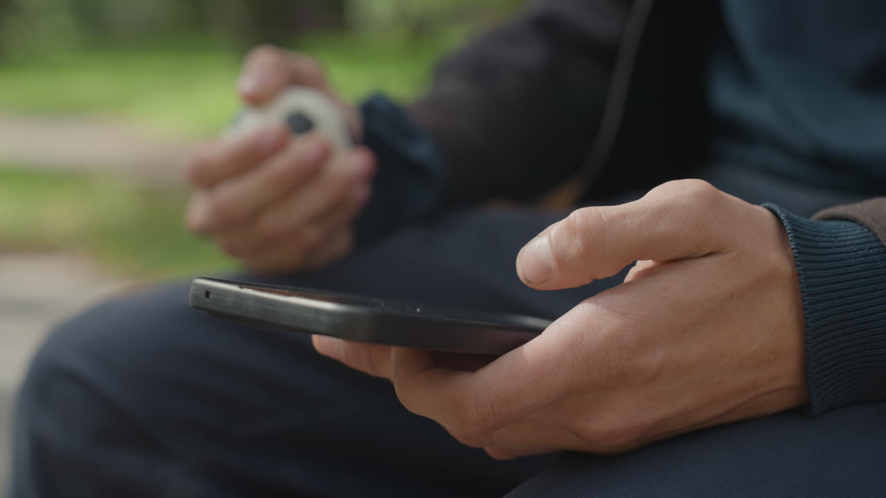 closeup of man using phone outdoors with small soccer ball in other hand, bench and grass background, warm sunlight and shallow depth creating relaxed trainingbreak mood thumb scrolling