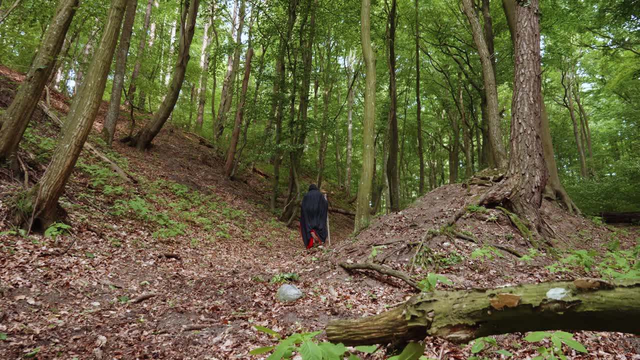 숲 속을 걷고 있는 낫을 움켜쥐고 검은 두건을 쓴 망토를 입은 잔인 사신의 뒷모습