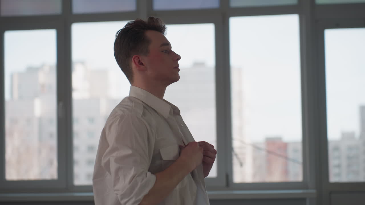 side view of young man adjusting white shirt with focused expression in bright room, standing near large window with blurred urban background, casual movement during quiet indoor moment