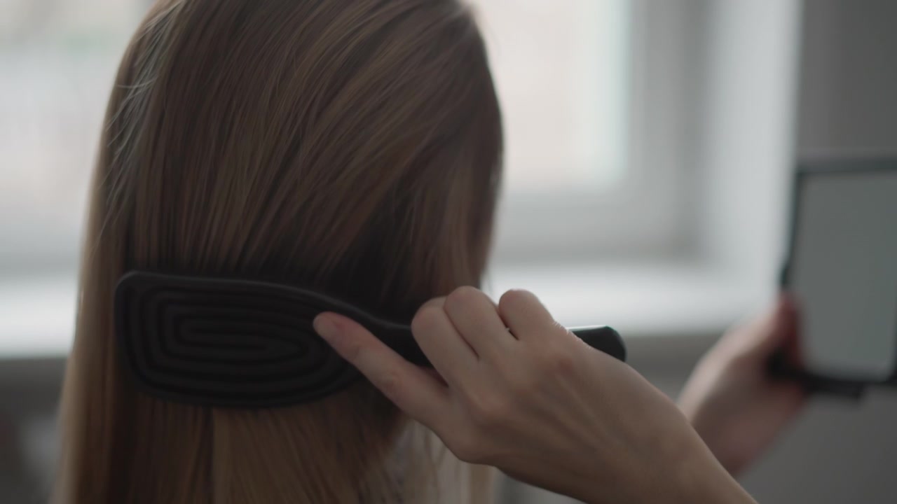 Close up of woman brushing long brown hair with smooth strokes while holding small mirror in one hand. action is performed indoors likely near window providing soft natural light focusing