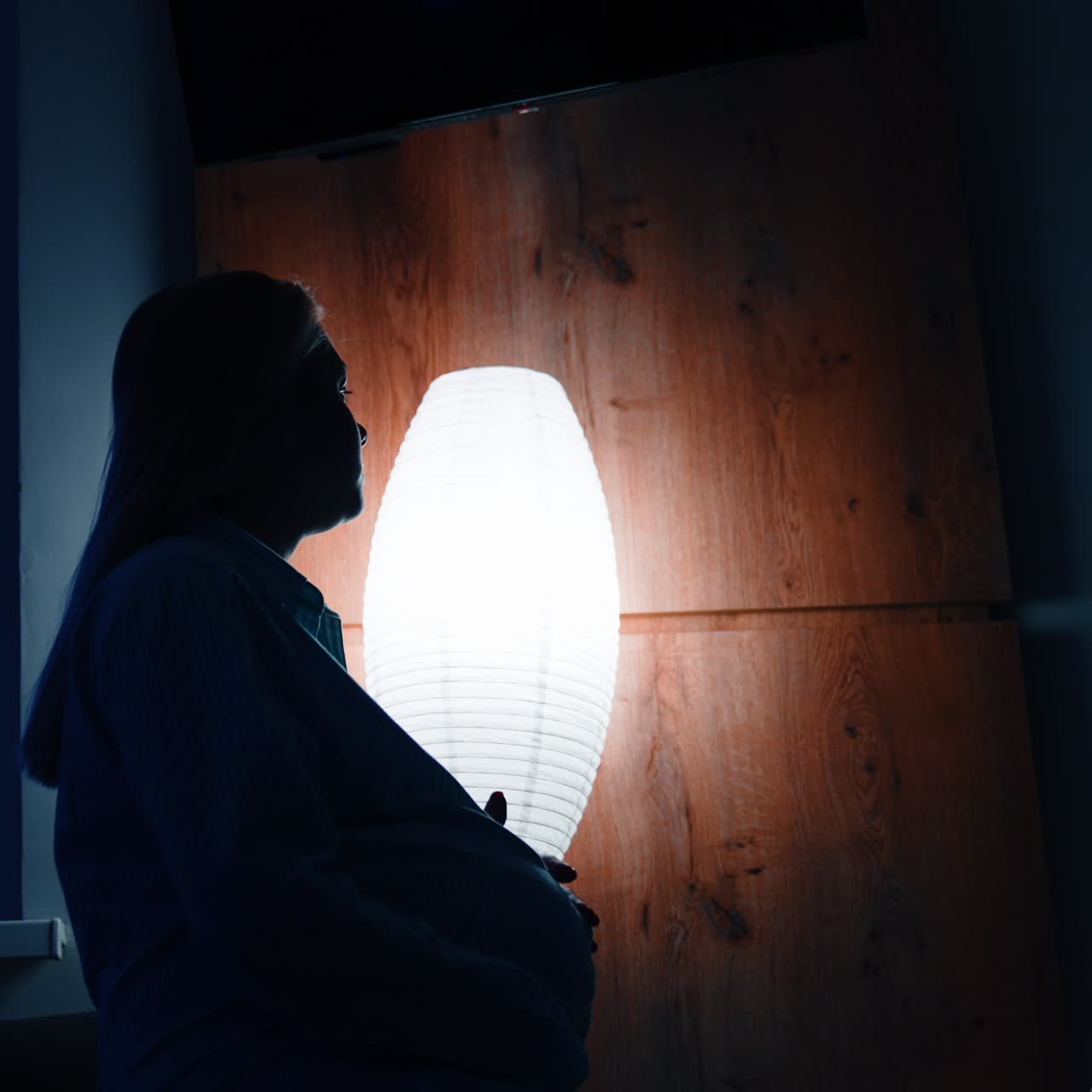 Silhouette of pregnant female sitting on the bed holding by the belly. Worried future mom in a dark room
