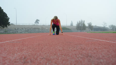 Vorderansicht-Einer-Kaukasischen-Sportlerin,-Die-Auf-Der-Laufstrecke-In-Einer-Sportstätte-Ihre-Startposition-Einnimmt,-4k