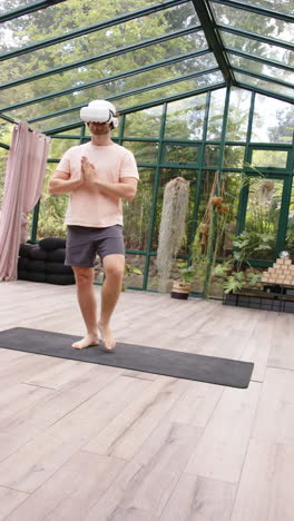 vertical video: practicing yoga, man wearing vr headset balancing on one leg in glass house
