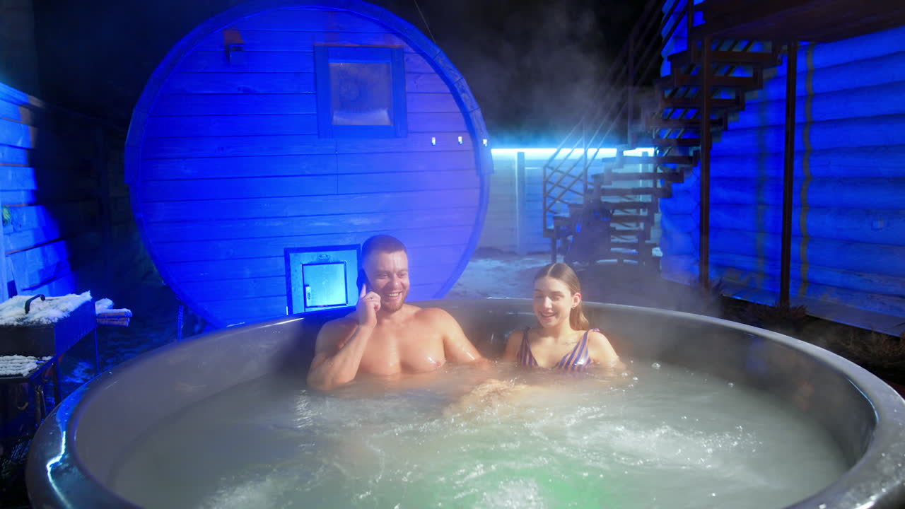 Caucasian man speaking on the phone and woman in stripy swimsuit sit in hot tub. Relax and spa concept.