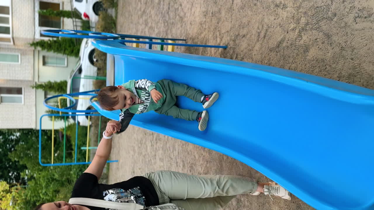 Active energetic Caucasian baby boy sits on the slide waving feet. Kid slides down supported by mom. Vertical screen.
