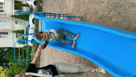 Active energetic Caucasian baby boy sits on the slide waving feet. Kid slides down supported by mom. Vertical screen.
