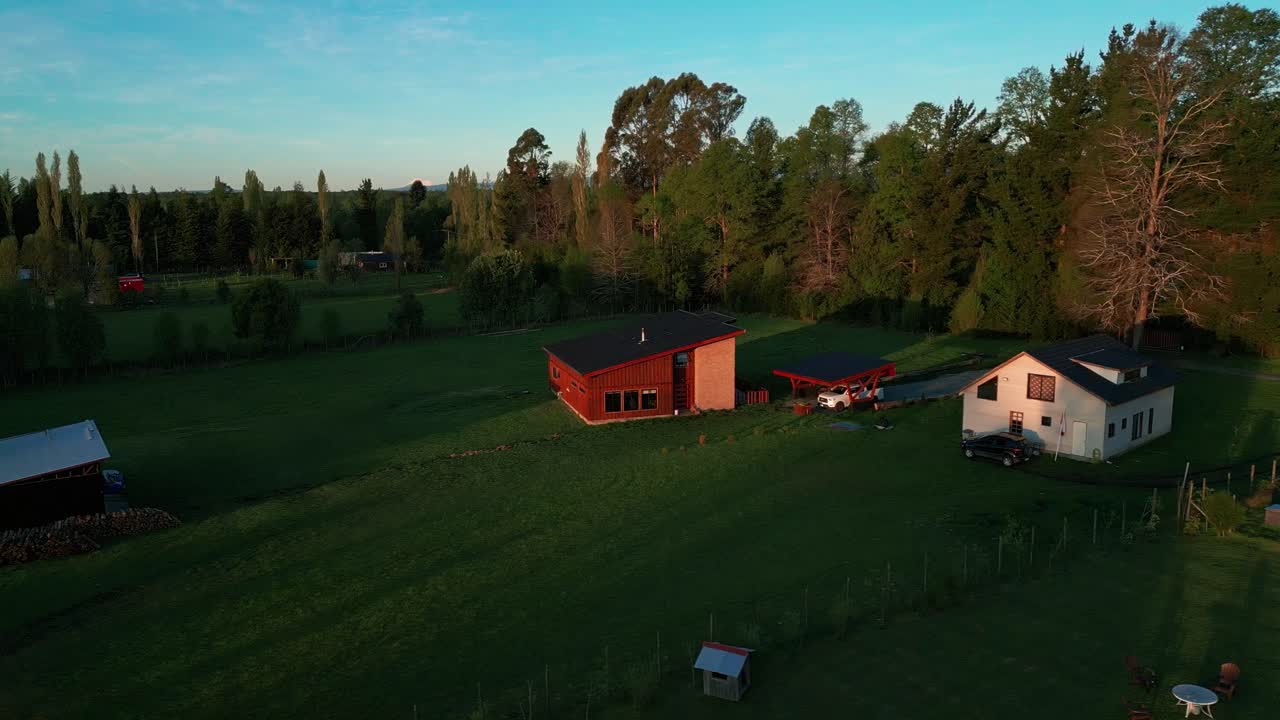 Drönarvy över ett lugnt lantligt landskap vid gyllene timmen, med två hus, frodig grönska och omgivande träd, som framkallar lugn och naturkänsla