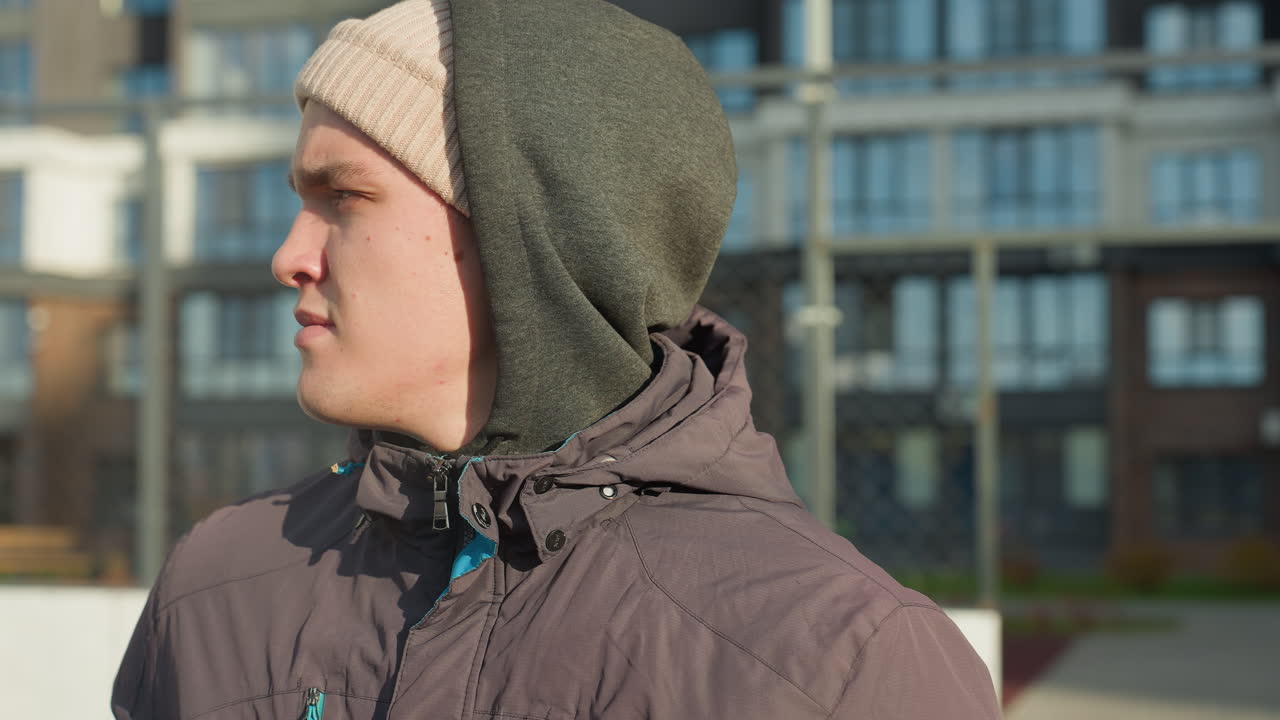 후디와 베니를 입은 어린 소년의 클로즈업은 축구장 위에 서서 집중된 표정으로 주변을 바라보고, 높은 주거 건물과 배경에 네트, 빛이 비추는 장면