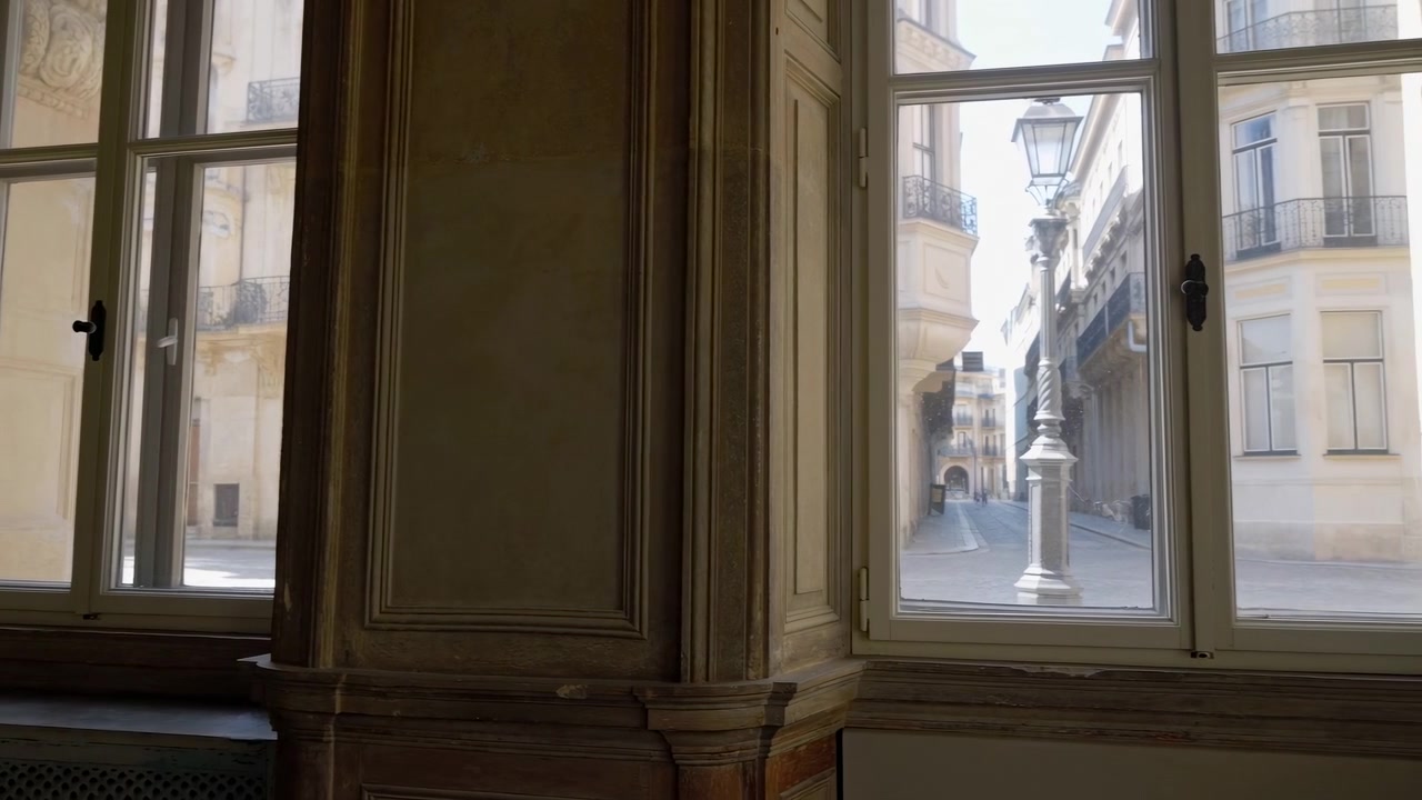 La calda luce del giorno riempie l'interno di un vecchio edificio mentre porte in legno a pannelli aperte e grandi finestre incorniciano una tranquilla strada cittadina, una scalinata e una facciata esterna sotto una morbida luce naturale e ombre.