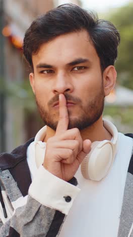 Serious indian young man presses index finger to lips makes silence hush gesture in city street