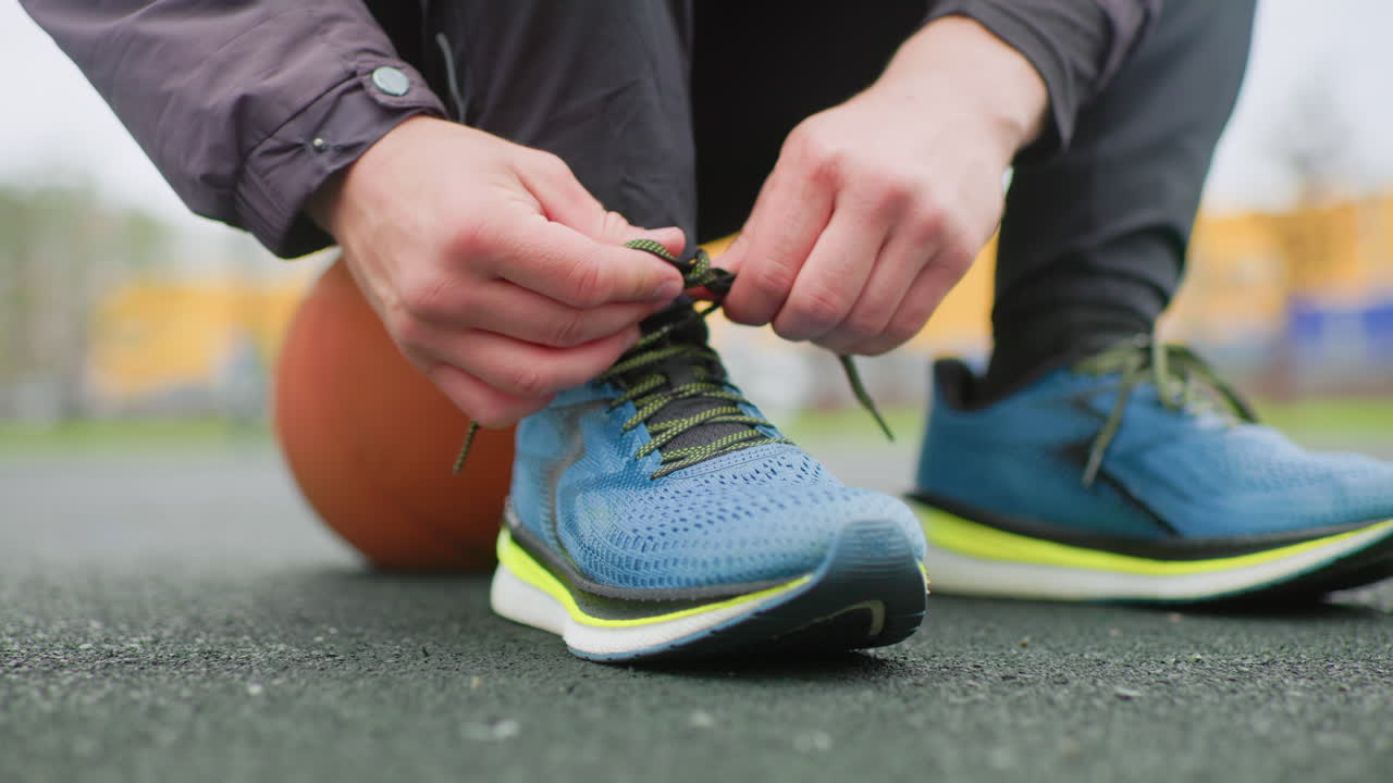 Atleta amarrando tênis de corrida azul, close-up das mãos dando nó nos cadarços com uma bola de basquete sob o joelho em uma quadra de asfalto ao ar livre, luz da manhã, jaqueta e leggings casuais, aquecimento pré-treino, focado