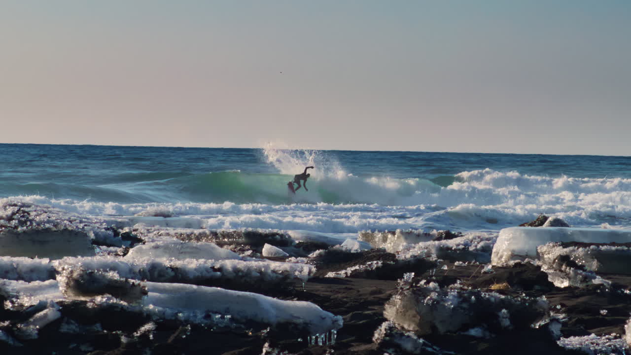 Onde oceaniche che si infrangono lungo la costa rocciosa con blocchi di ghiaccio galleggianti nel mare freddo, mentre un surfista esegue un cutsback al rallentatore, panoramica lenta a sinistra.