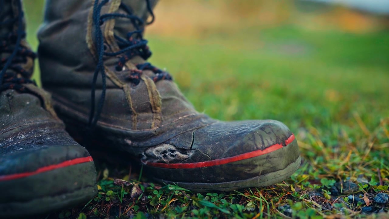 Sepasang Sepatu Bot Kerja yang Sangat Usang Berdiri di Tanah Berumput, Menyoroti Tanda-tanda Pekerjaan Luar Ruangan yang Ekstensif, Penggunaan Kasar, dan Ketahanan di Lingkungan Kerja Pedesaan - Bidikan Jarak Dekat