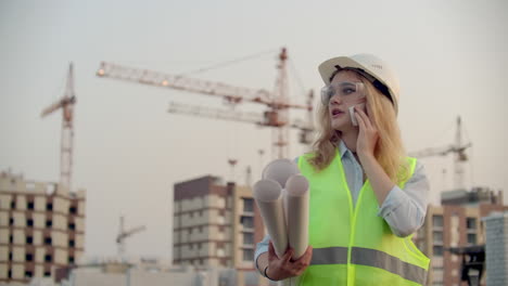 mujer constructor hablando por teléfono con el contratista con dibujos en sus manos en el fondo de los edificios en construcción y grúas