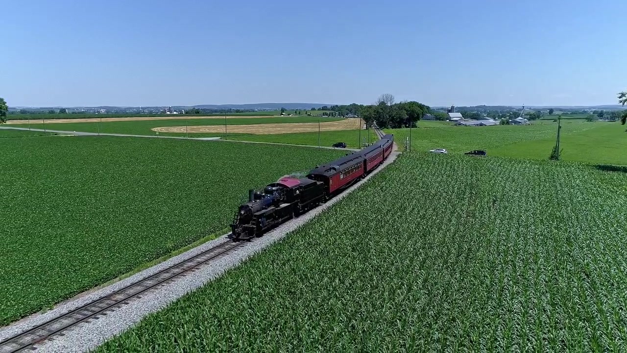 Un treno a vapore sbuffa lungo i suoi binari, circondato da campi verdi e coltivazioni in Pennsylvania in una limpida giornata estiva. Paesaggi panoramici creano uno sfondo nostalgico per questo viaggio classico.