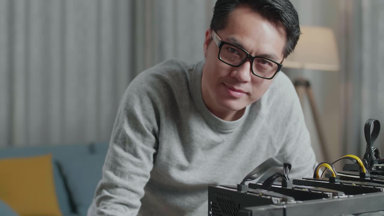미소 짓는 아시아 남자가 안경을 쓰고 엄지손가락을 올려놓고 노트북과 광산 기계를 사용하여 암호화폐를 채굴하는 제스처를 보여줍니다.
