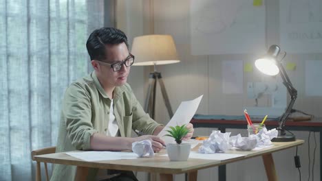 화가 난 아시아 남자 가방 디자이너는 사무실에서 레이아웃 본드를 보면서 머리를 흔들고 있습니다.