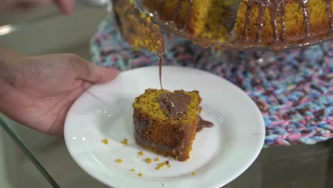 serving a simple homemade carrot cake with chocolate sauce on a white plate with a metal spatula lifting the piece of candy with the syrup dripping onto the plate