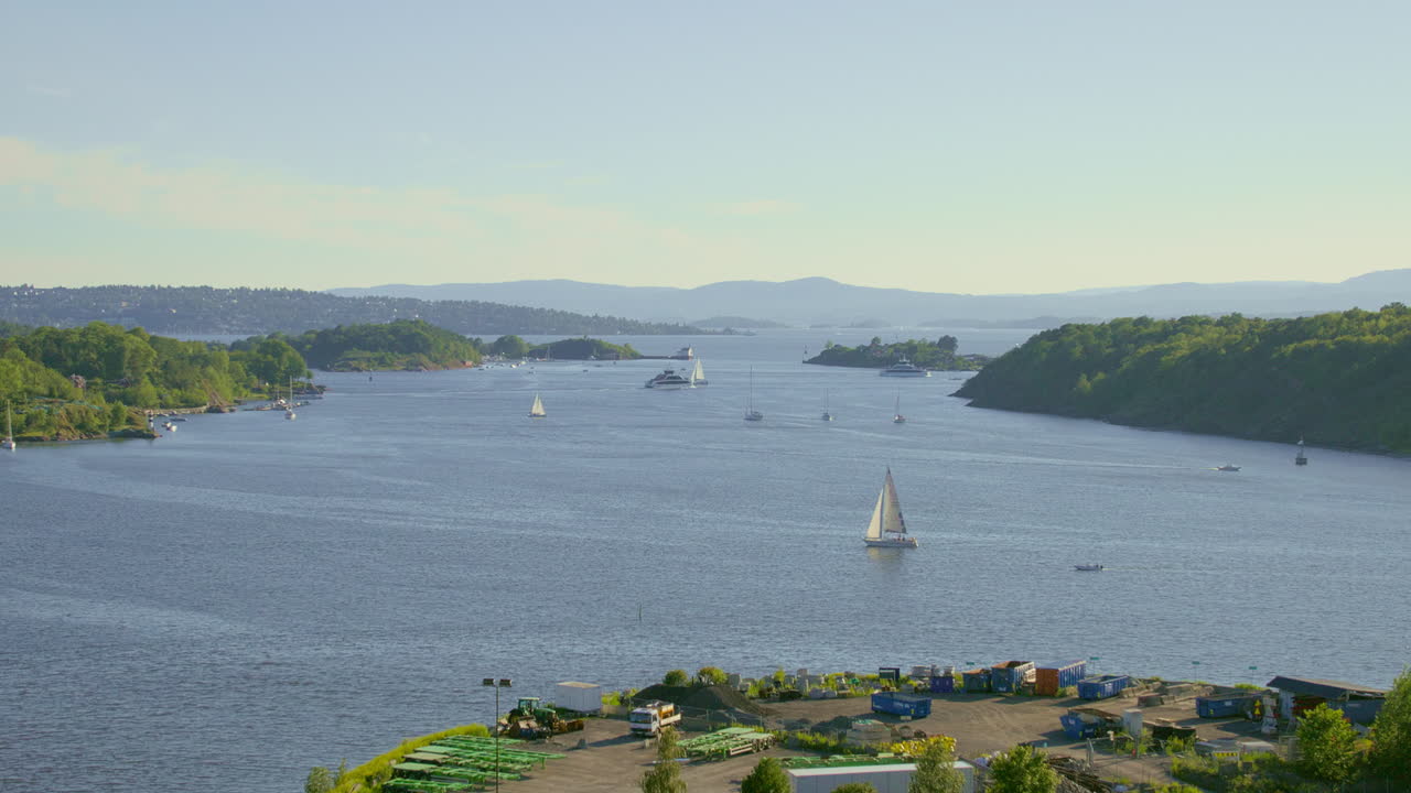Sailboats and yachts in a tranquil bay with green hills and islands under a clear blue sky.