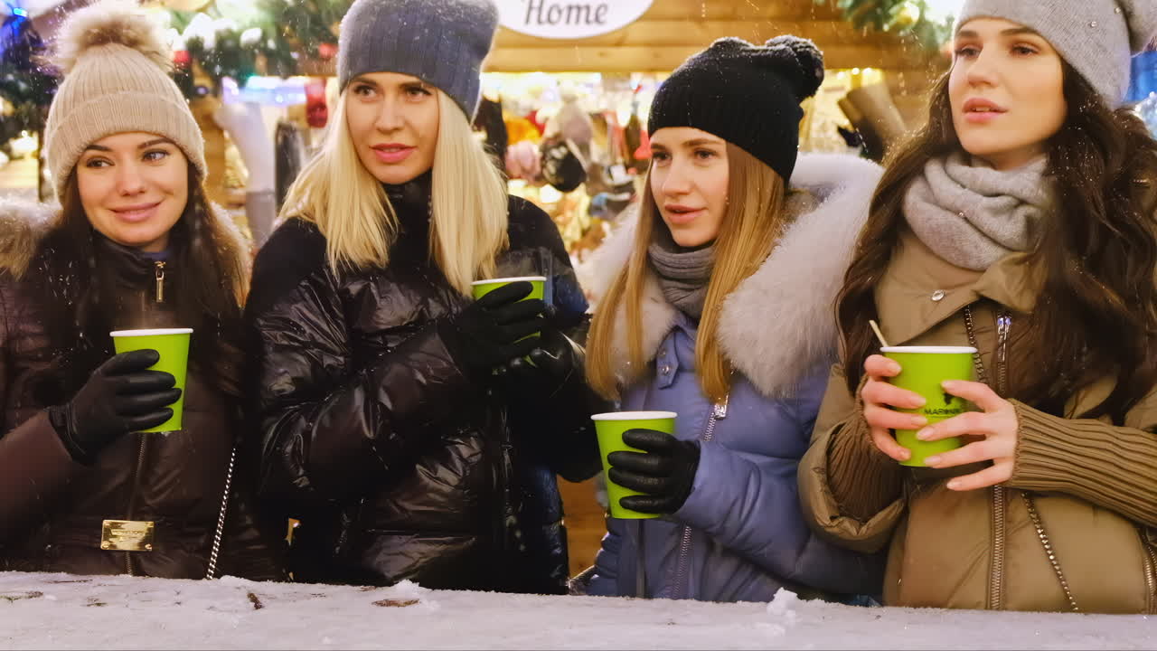 Four friends gather at a winter market, sipping warm drinks and laughing. They are dressed in cozy hats and jackets under festive lights. The atmosphere is joyful and vibrant
