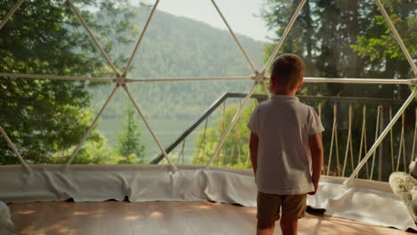 child walks to glass dome at glamping. little barefoot boy looks through clear wall on hotel terrace. kid enjoys nature beauty on summer holiday