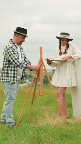 a smiling woman stands in a grassy field with her husband. the man, wearing a black hat and plaid shirt, sets up an easel while the woman, dressed in a white dress and black hat, holds a paint palette