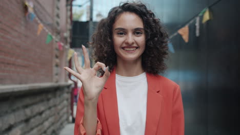 smiling woman in red blazer giving ok sign in a city alley