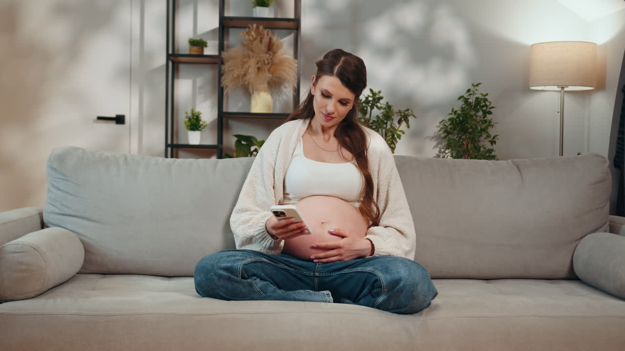 Expecting mother sits crosslegged on sofa holding phone and touching belly relaxed indoor setting