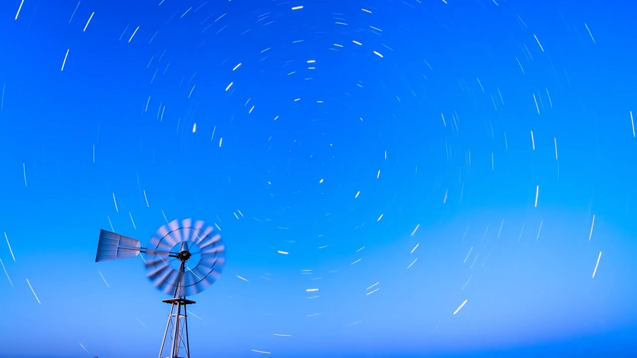 回転する風車の羽根、地球の自転が平野に星の軌跡を描き、地平線が輝く。コピースペースあり。