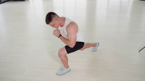 White man stretching body in tiled gym house on light wooden floor performing side lunge with hands clasped wearing white tank top black shorts and light sneakers under bright windows