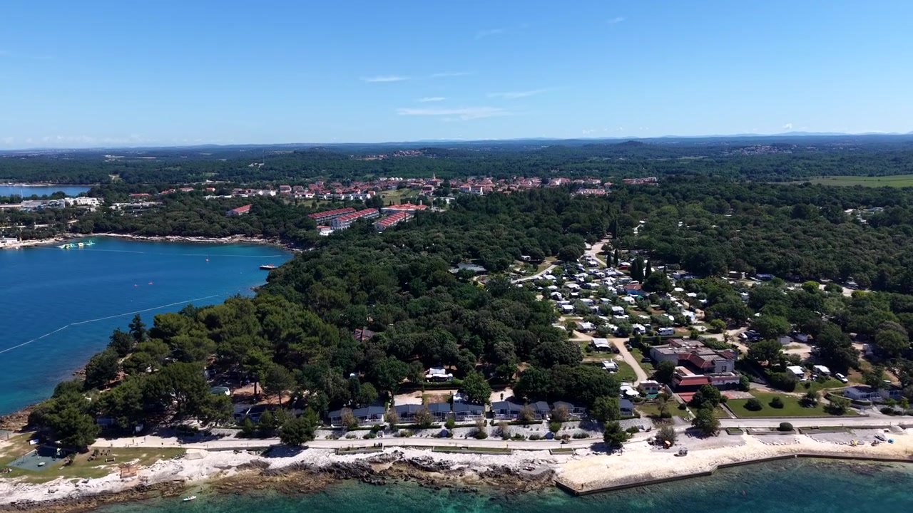 Furuomkranset kystpromenade snor seg langs steinete kystlinje, campingvogner sprer seg i skogen mens en historisk kystby brer seg utover en rolig turkis Adriaterhavsbukt. Kroatia. Luftfoto med drone som stiger opp (vidvinkel).