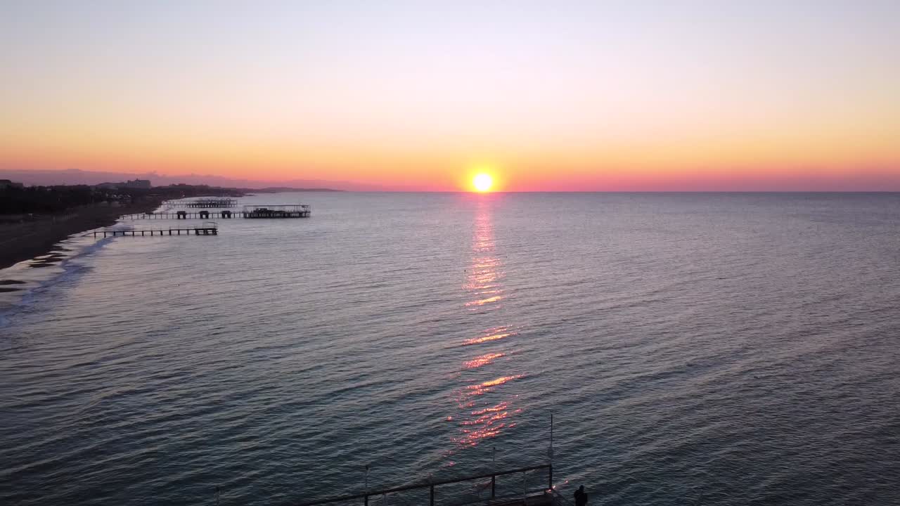 Man standing on an old pier, sunrise on the horizon, wavy sea aerial shot