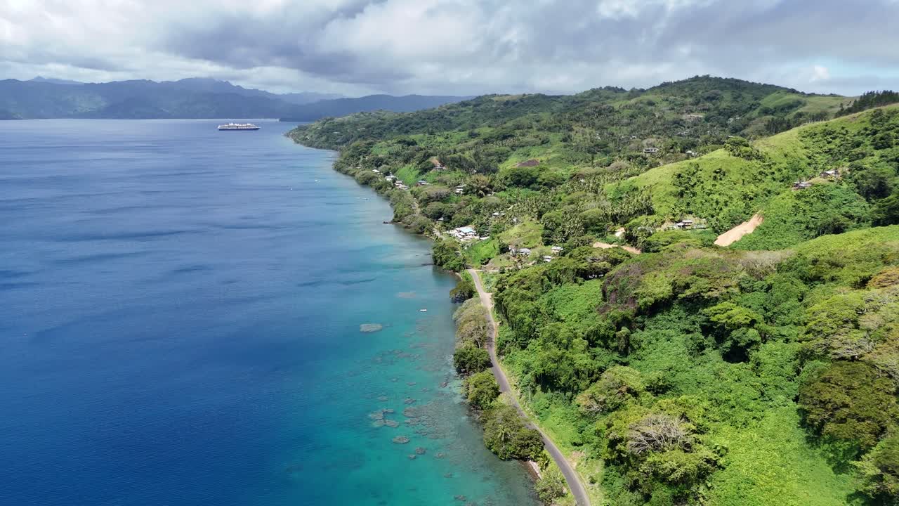 Drone-opptak i kinokvalitet med utsikt over frodige, tropiske fjell og det krystallklare, blå vannet rundt en avsidesliggende øy i Fiji-arkipelet på en lys og solfylt dag