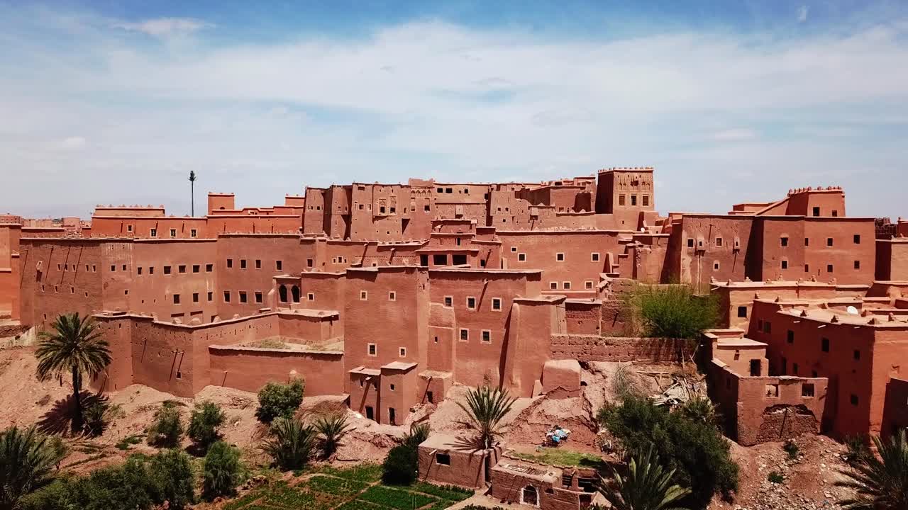 Amplia vista panorámica de una kasbah marroquí en Ouarzazate con vistas a palmeras y tierras de oasis cultivadas. Fuerte contraste entre la arquitectura desértica y la vegetación, ideal para imágenes de viajes y cultura
