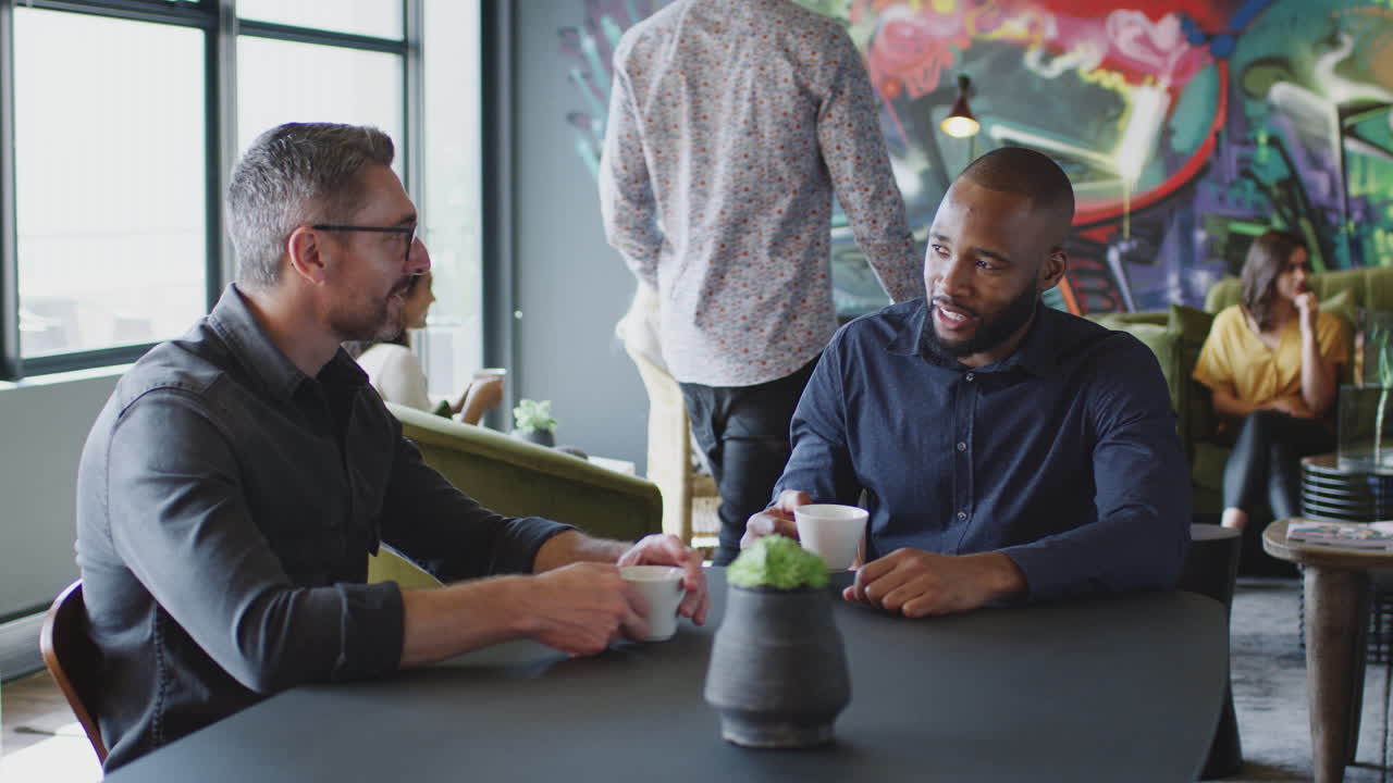 Two male creative colleagues talking in their workplace cafe, three quarter length, arc shot