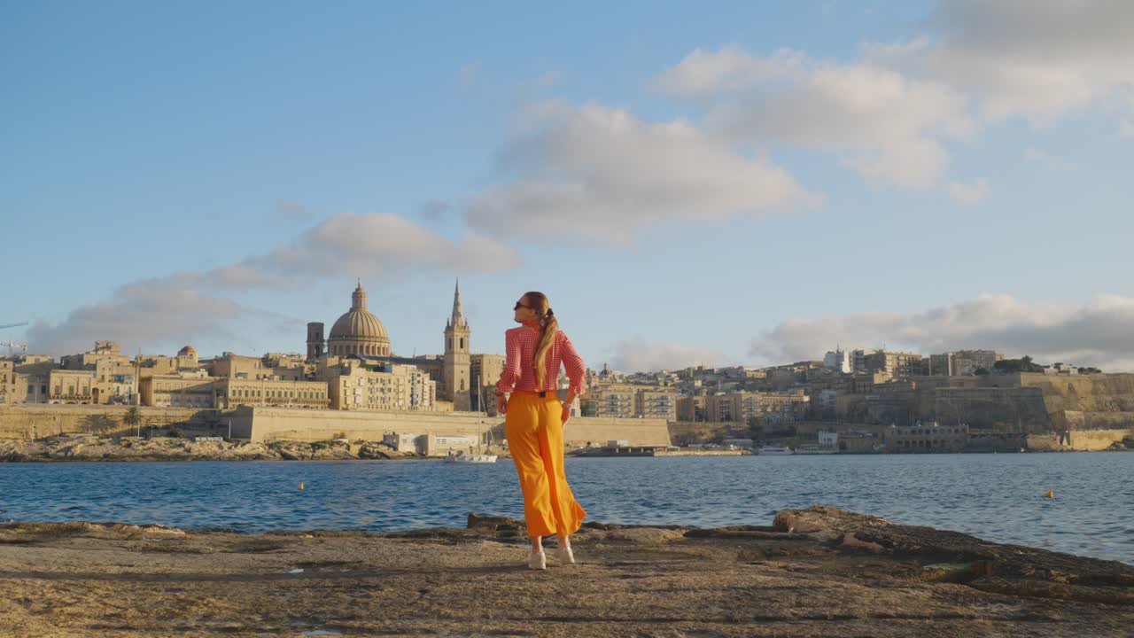 Vista posteriore di una donna che cammina in riva al mare verso lo skyline e l'architettura storica di La Valletta.