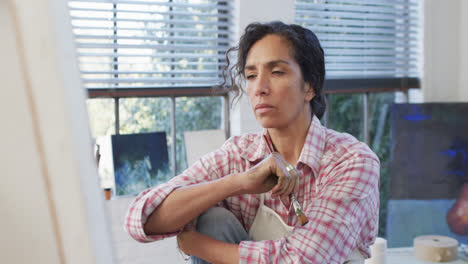 Biracial-female-artist-in-apron-holding-brush-and-looking-at-canvas-in-studio,-slow-motion