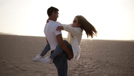 Adorable-adult-couple-in-white-shirts-and-jeans-man-holding-her-woman-on-arms-and-turning-her-around,-have-fun-emotionally,-laugh,smile-on-sunset-at-desert-crazy-in-love,-emotions-and-relationship.-Whirling-around