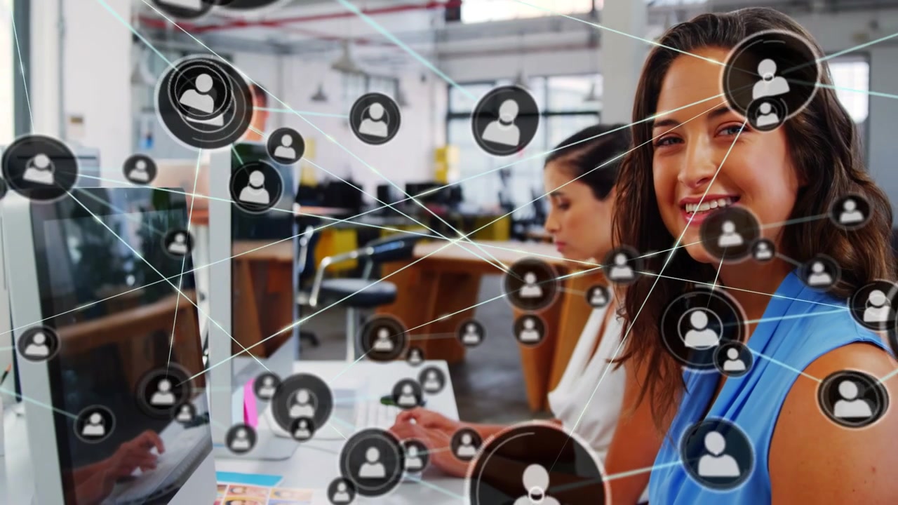 Female coworkers typing while monitor spawning user-icon network, visualizing team sync for tech
