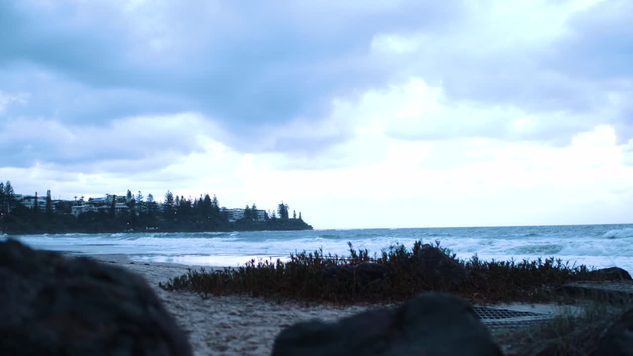 A gorgeous beach sits infront of rocks and foliage crashing its waves onto the wet and coarse sand as the sky paints a blue tint in its fluffy clouds from the descending sun