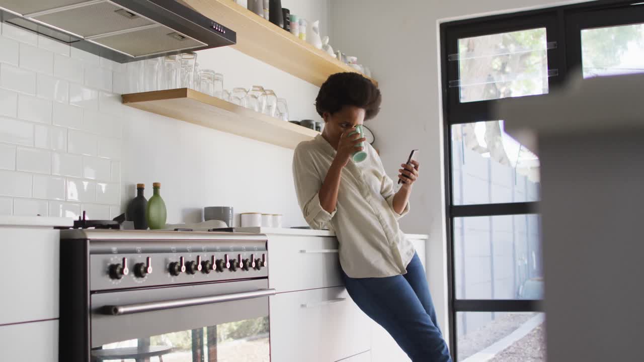 Donna afroamericana felice che beve caffè e usa lo smartphone in cucina