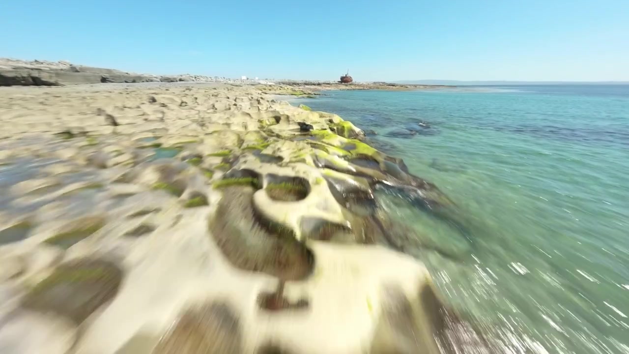 Una telecamera FPV sfreccia bassa lungo la costa calcarea fessurata e l'acqua turchese brillante, seguendo il relitto lontano della Plassey.