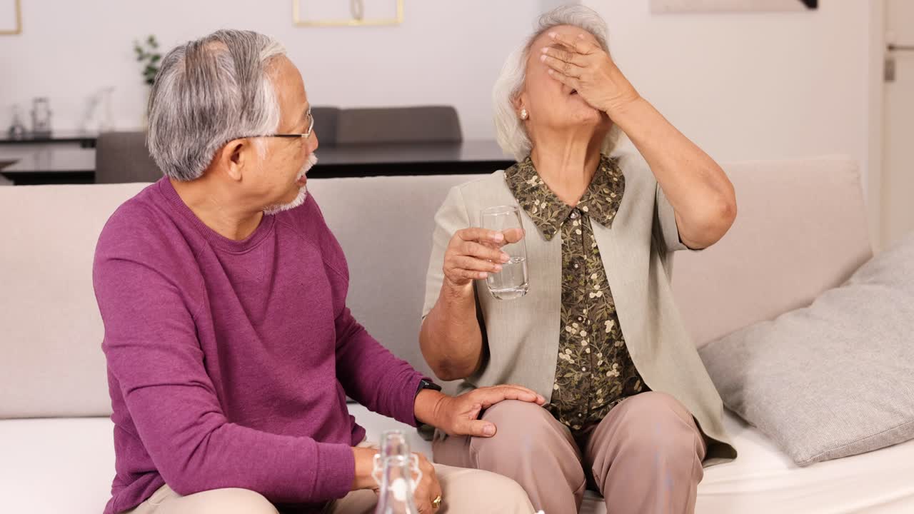 An elderly Asian couple sits on a sofa in a bright living room as the woman takes medication with water, supported by her partner. Natural lighting, steady camera, warm and caring mood