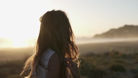 Slow-motion-handheld-shot-of-a-young-beautiful-woman-dressed-in-white-beach-stroking-through-her-hair-standing-on-the-sandy-beach-with-the-sea-in-the-background-during-a-beautiful-sunset