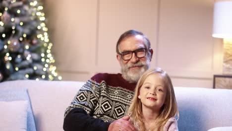 close up portrait of cheerful family old grandpa with cute small girl sitting in decorated room near xmas tree smiling and hugging on new year's eve