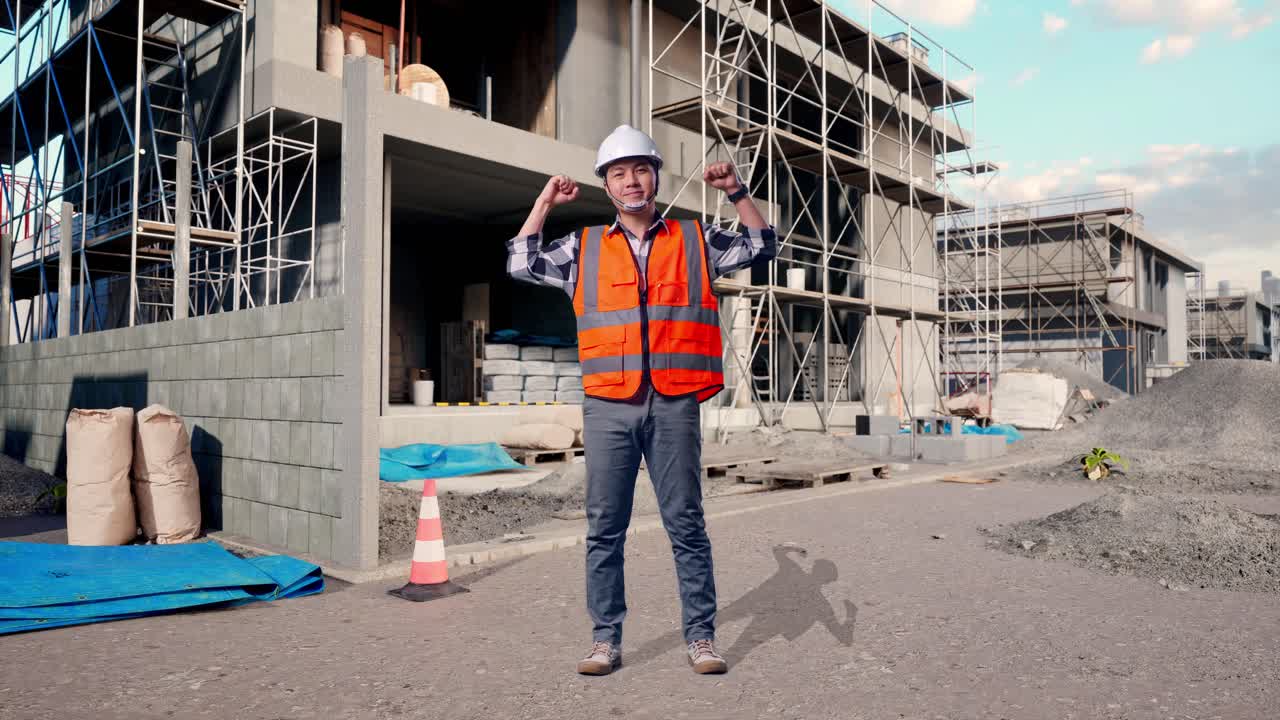 Full Body Of Asian Male Engineer With Safety Helmet Flexing His Bicep And Smiling To Camera While Standing At Building & Construction Site in Progress to New House